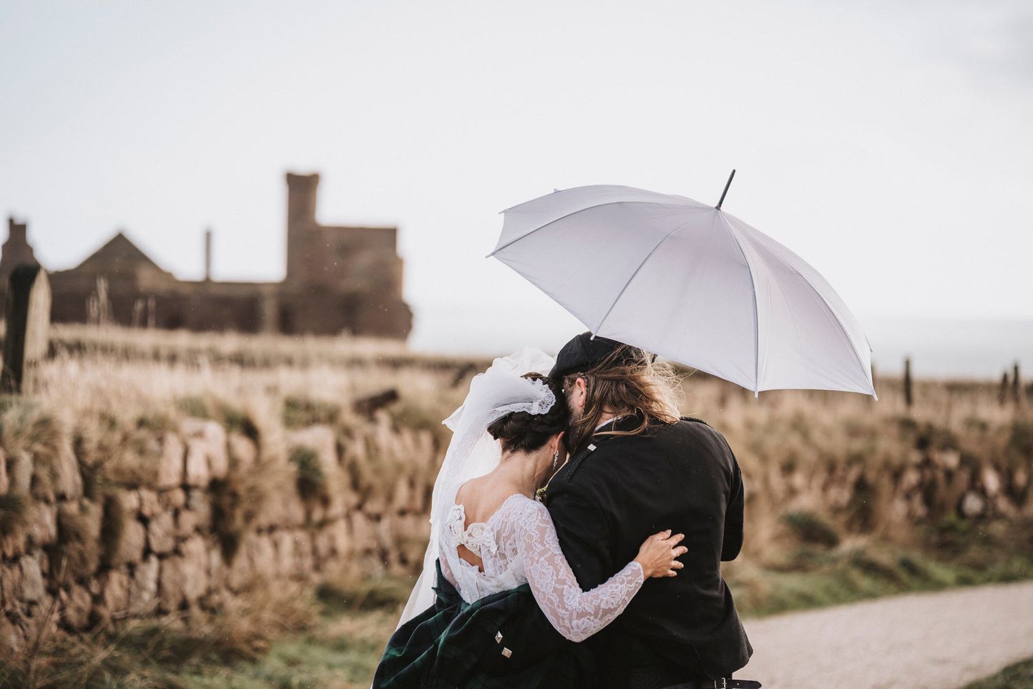A couple shares an intimate moment under a white umbrella near a stone wall and historic building.