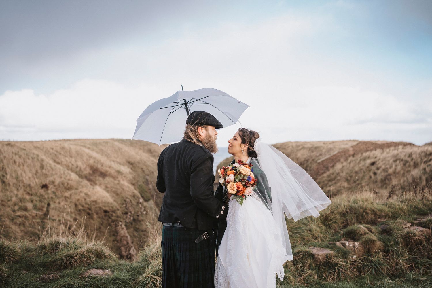 A couple shares a romantic kiss on a scenic hilltop overlooking a rugged coastal landscape at sunset.