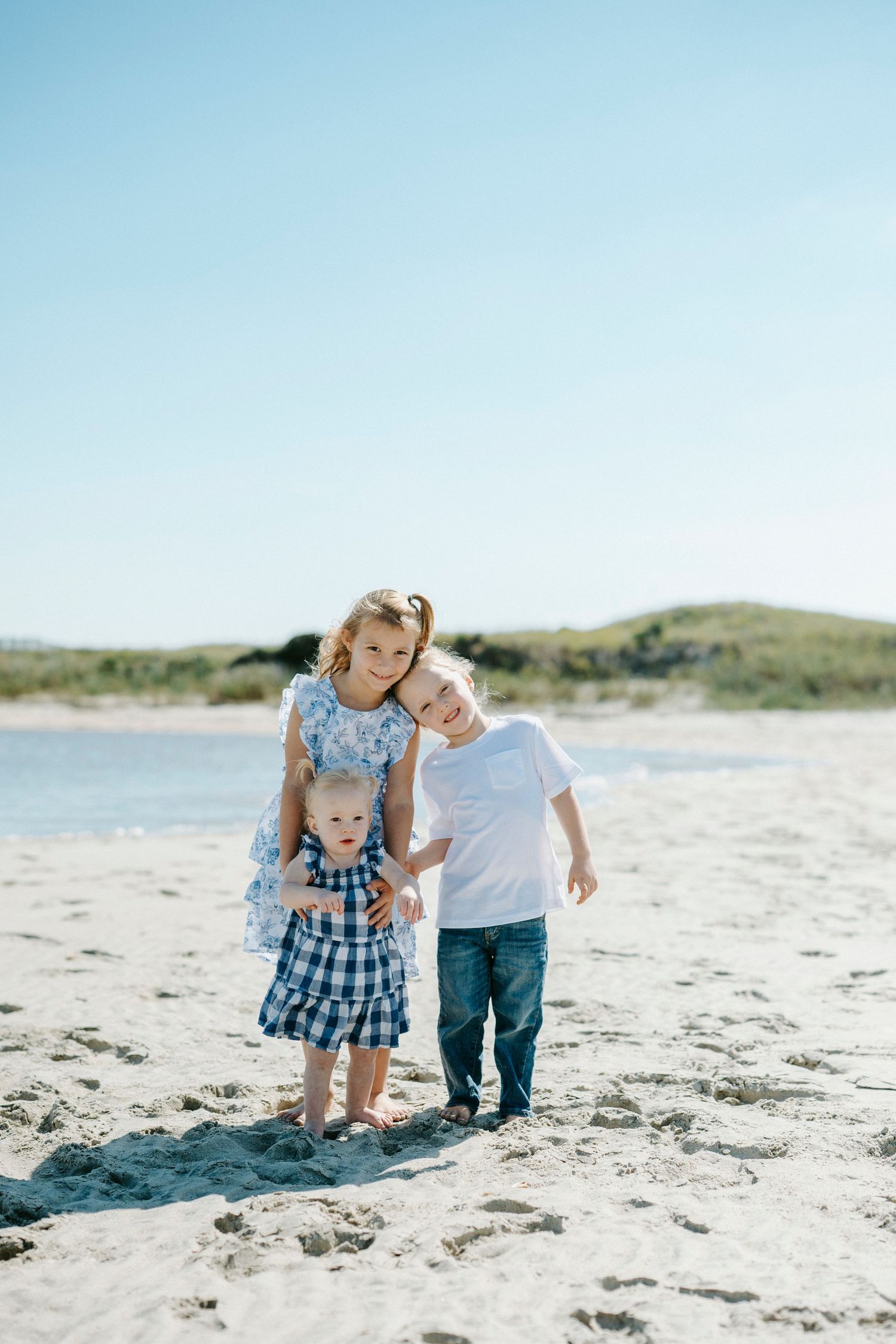 Two small children play together on a sunny beach with sand and dunes in the background.