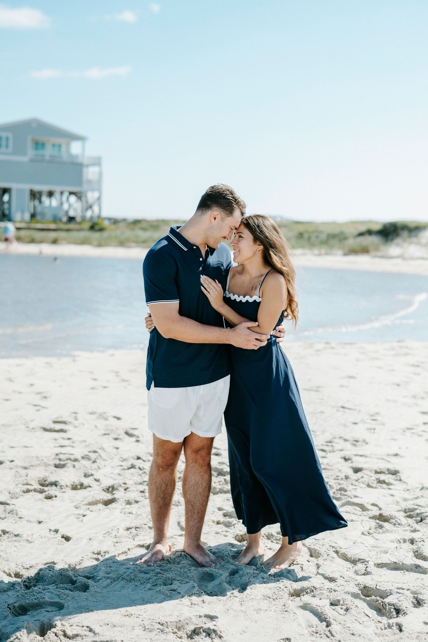 A romantic couple embraces on a sunny beach with coastal homes visible in the background.