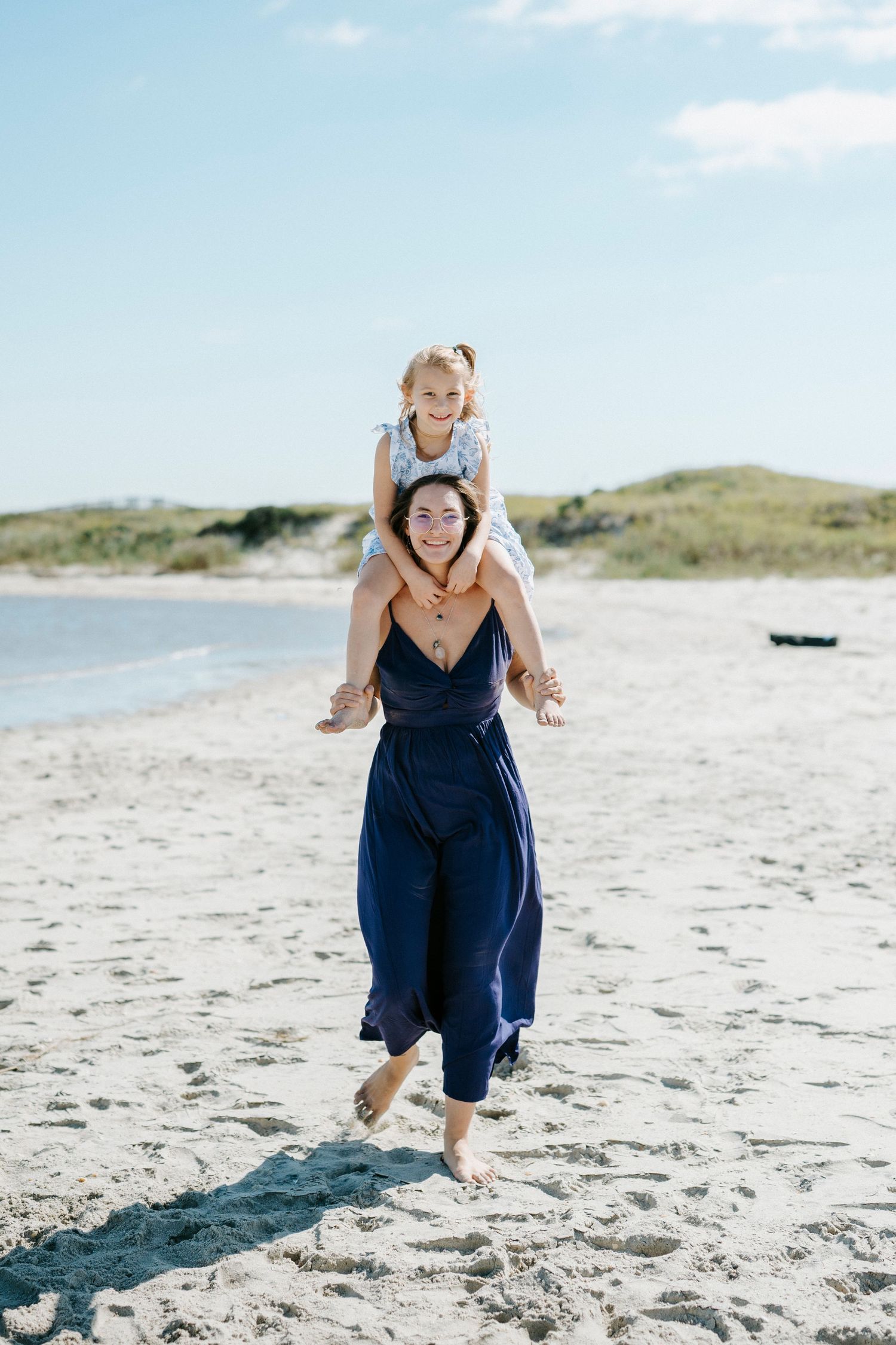 A person in a navy blue dress joyfully jumps and plays on a sunny beach with sand dunes in the background.
