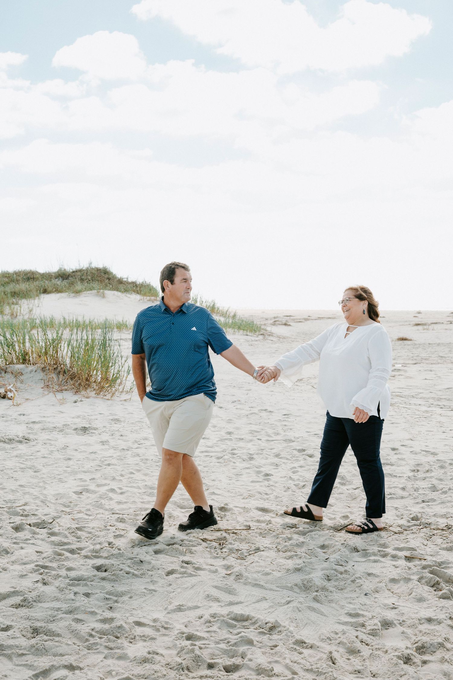 A couple playfully dances together on a sandy beach with dunes and sea grass in the background.