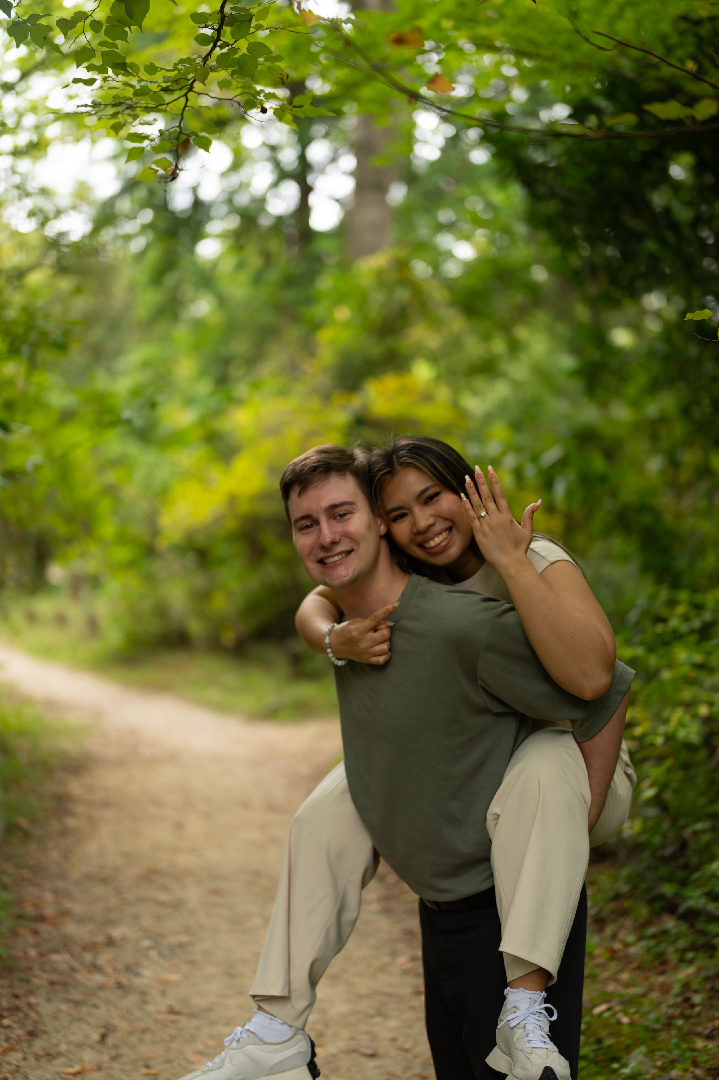 Happy couple posing on a woodland path, woman showing engagement ring on her hand.