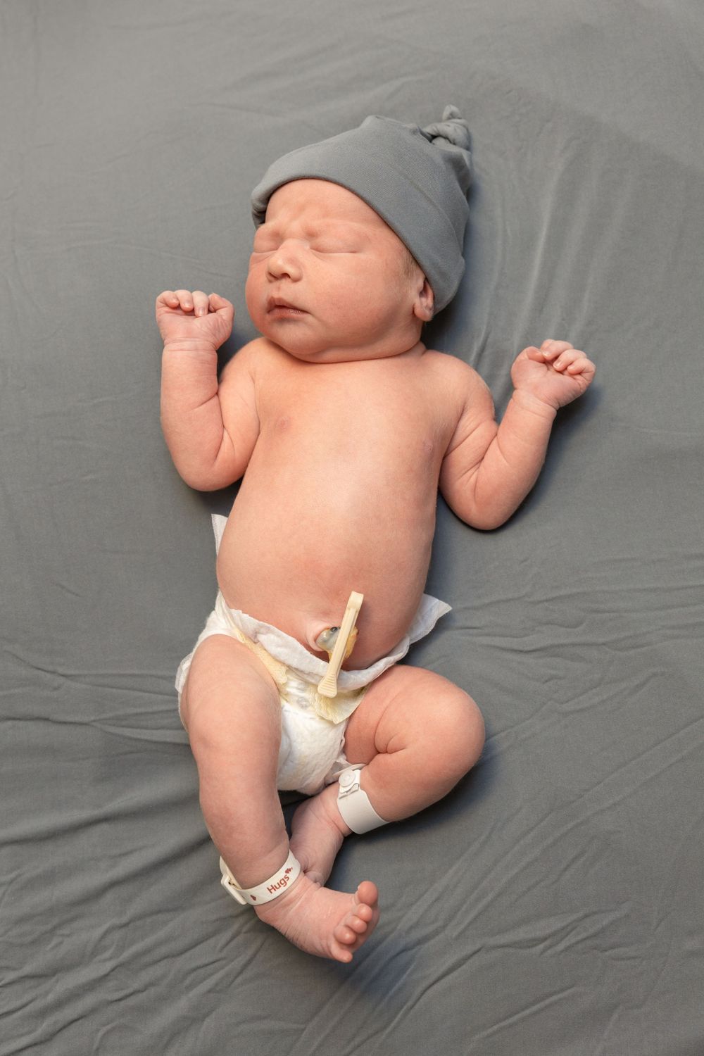 Newborn baby sleeping on gray sheets wearing a gray knot hat and diaper with umbilical cord clamp.