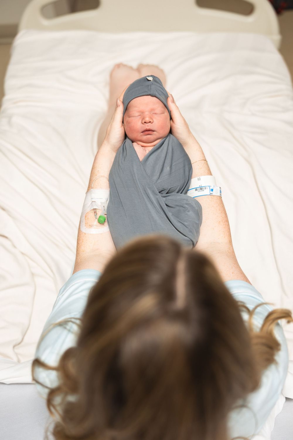 New mother in hospital bed holding swaddled newborn baby in gray wrap and hat after delivery.
