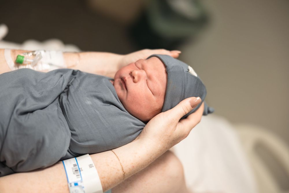 Newborn baby swaddled in gray blanket being held by parent wearing hospital wristband after delivery.