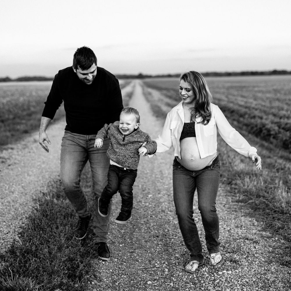 Black and white photo of a pregnant woman, man, and toddler walking on a rural path, joyfully swinging the child.