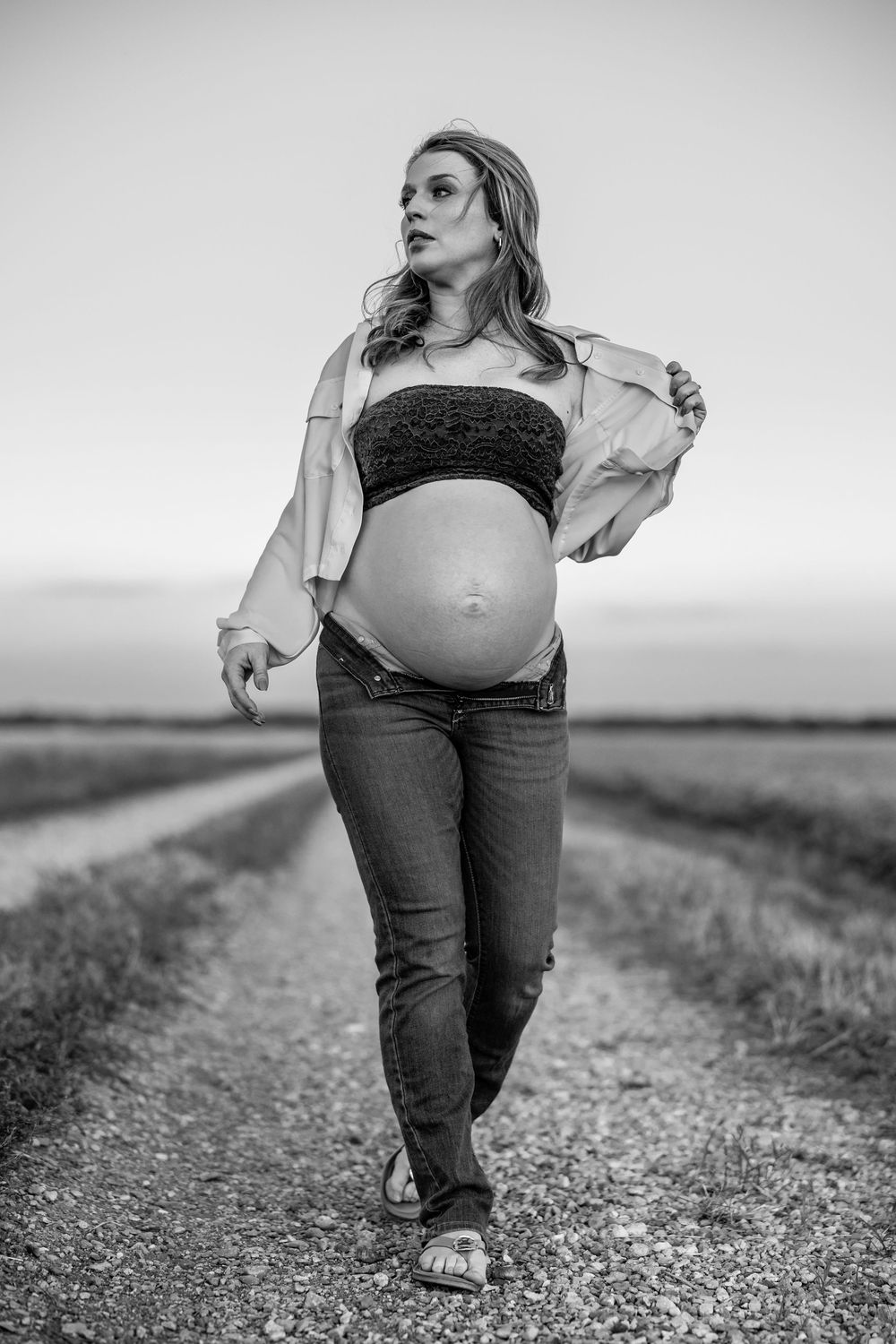 Black and white photo of a pregnant woman walking on a rural dirt road, showing her baby bump in jeans and open shirt.