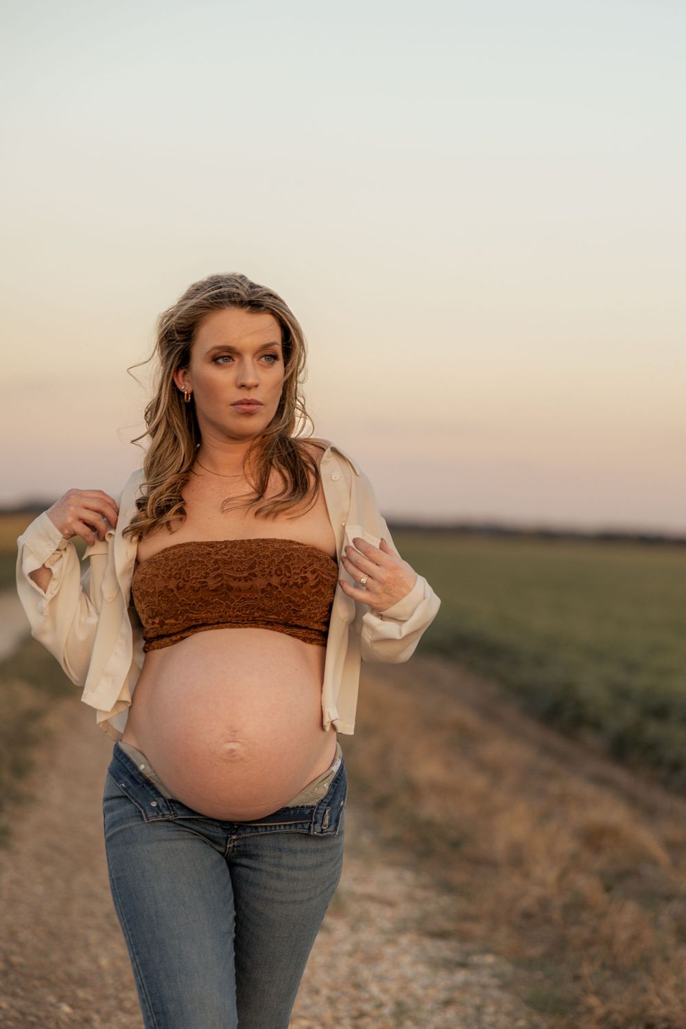 Pregnant woman in brown crop top and jeans poses on rural dirt road at sunset with baby bump exposed.