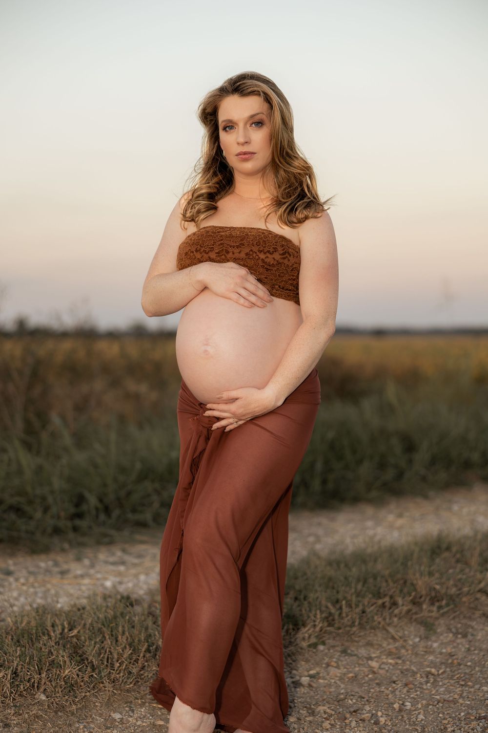 Pregnant woman in brown tube top and skirt cradling baby bump outdoors at sunset in a field.