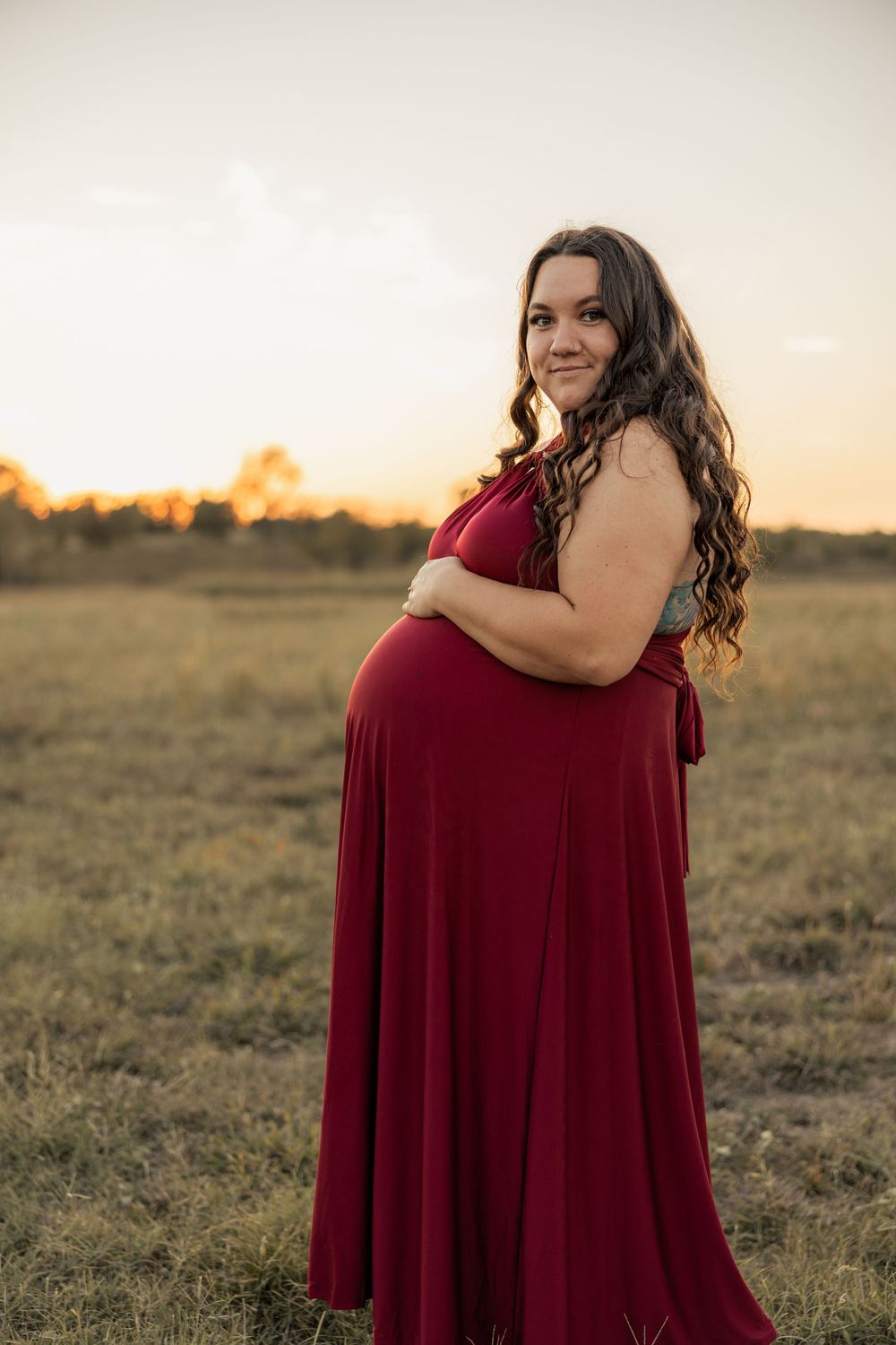 Pregnant woman in red maxi dress holding baby bump at golden hour in an open field.
