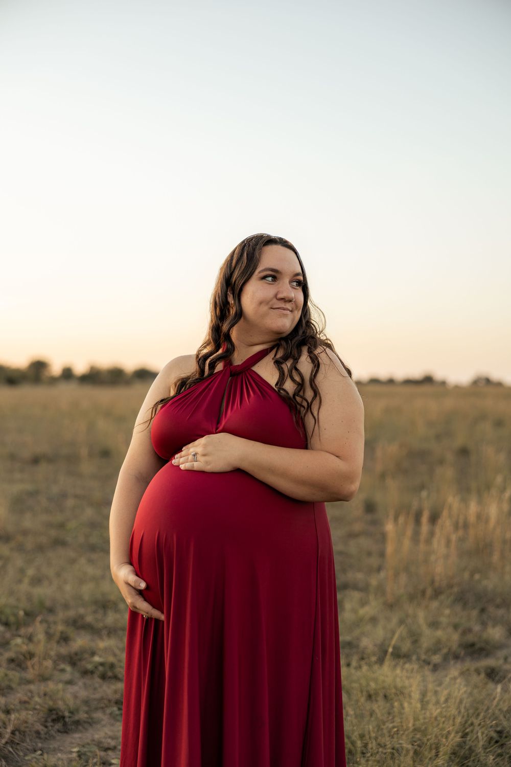 Pregnant woman in red halter dress cradling baby bump in golden field at sunset.