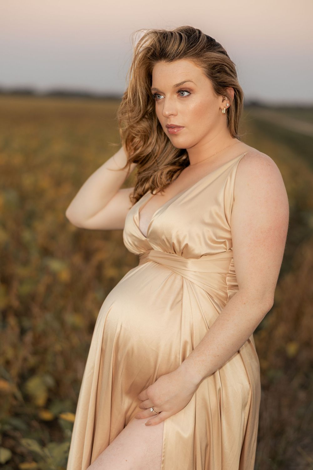 Pregnant woman in gold satin dress posing outdoors in a field at sunset, cradling her baby bump.