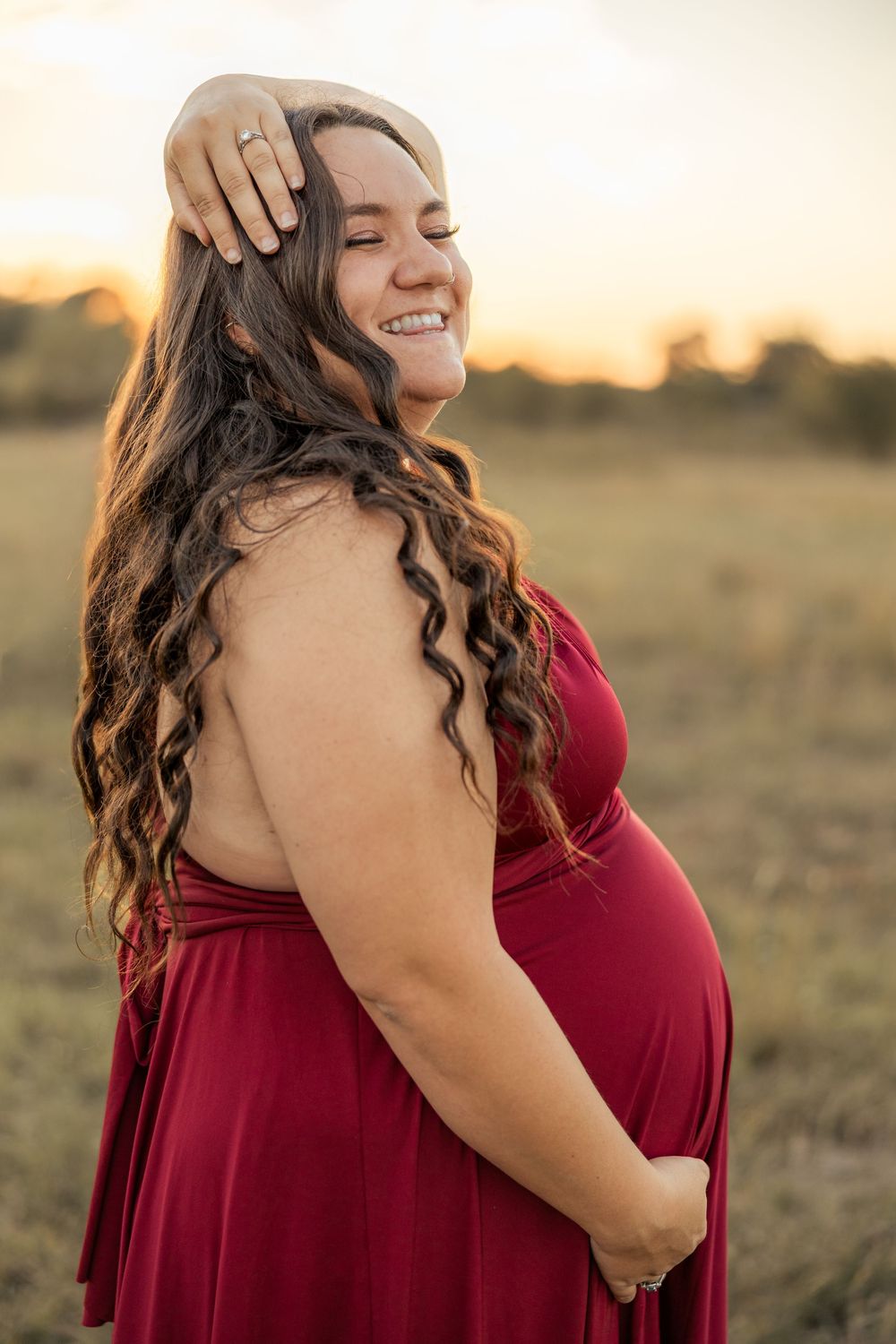 Smiling pregnant woman in red dress at golden hour, hand in hair, showcasing maternity glow in outdoor field setting.
