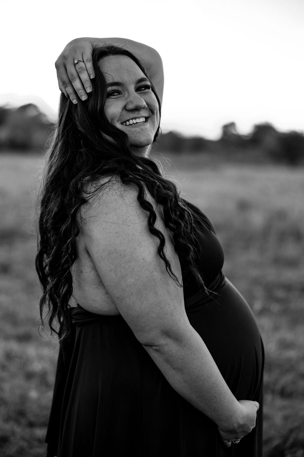 Smiling pregnant woman in black dress poses outdoors in a field, black and white maternity portrait photography.