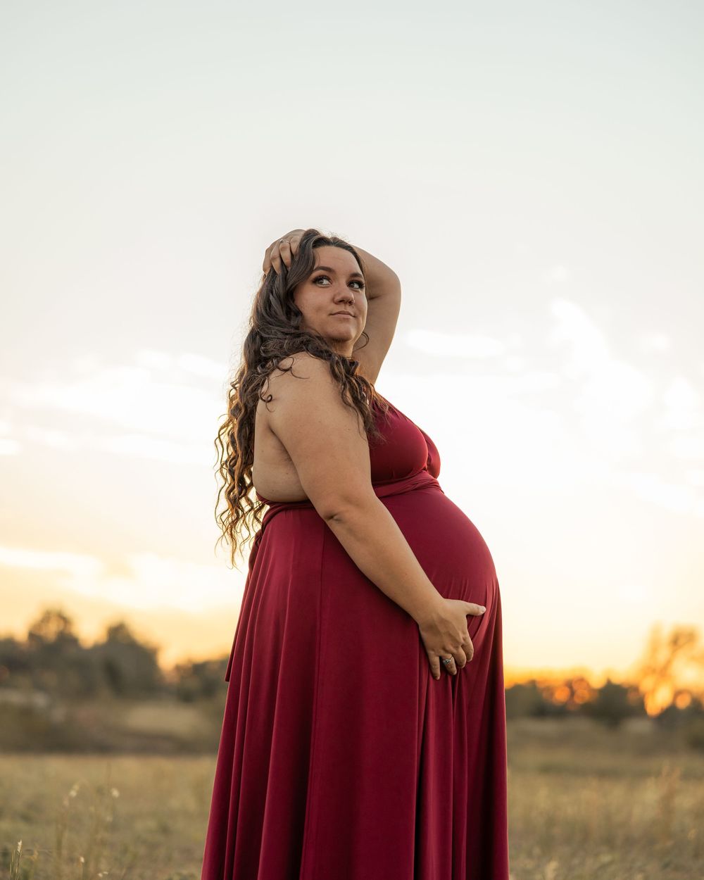 Pregnant woman in red gown poses at golden hour in a field, cradling her baby bump with long wavy hair.