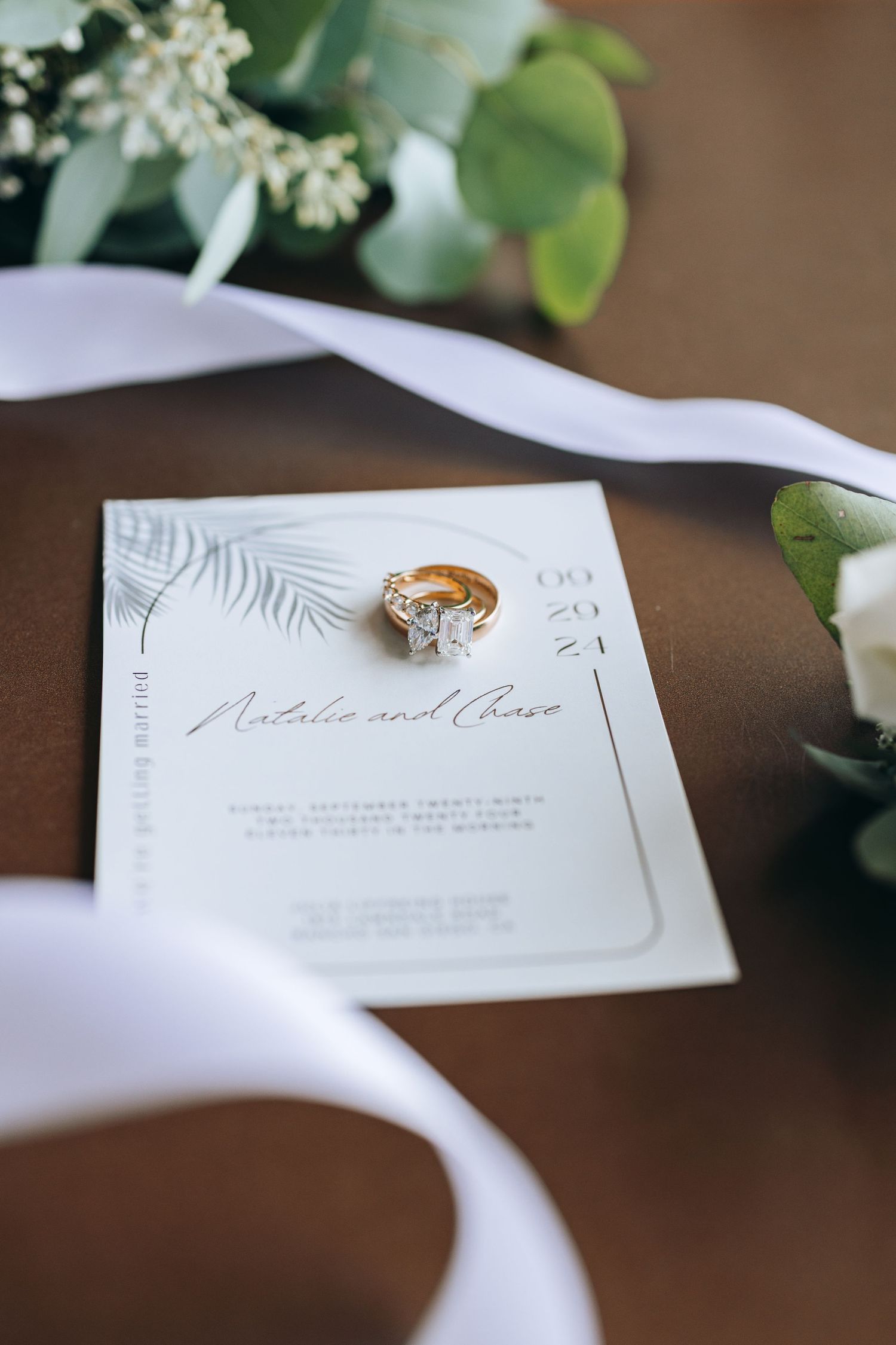 Elegant wedding invitation with gold ring photographed with eucalyptus and white ribbon.