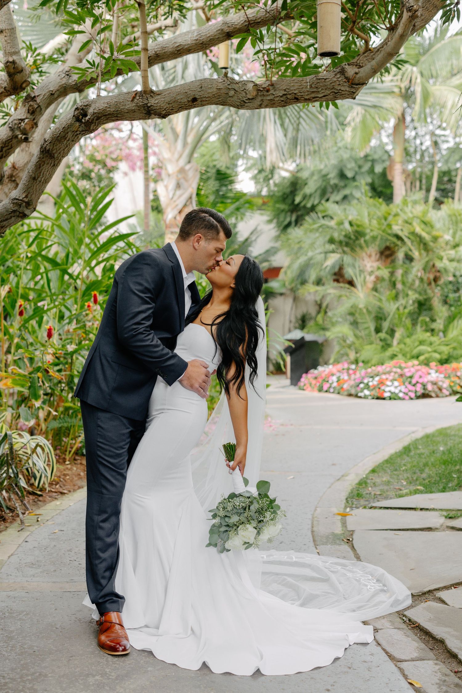 A couple shares a romantic kiss on a garden path surrounded by lush tropical plants and flowers.