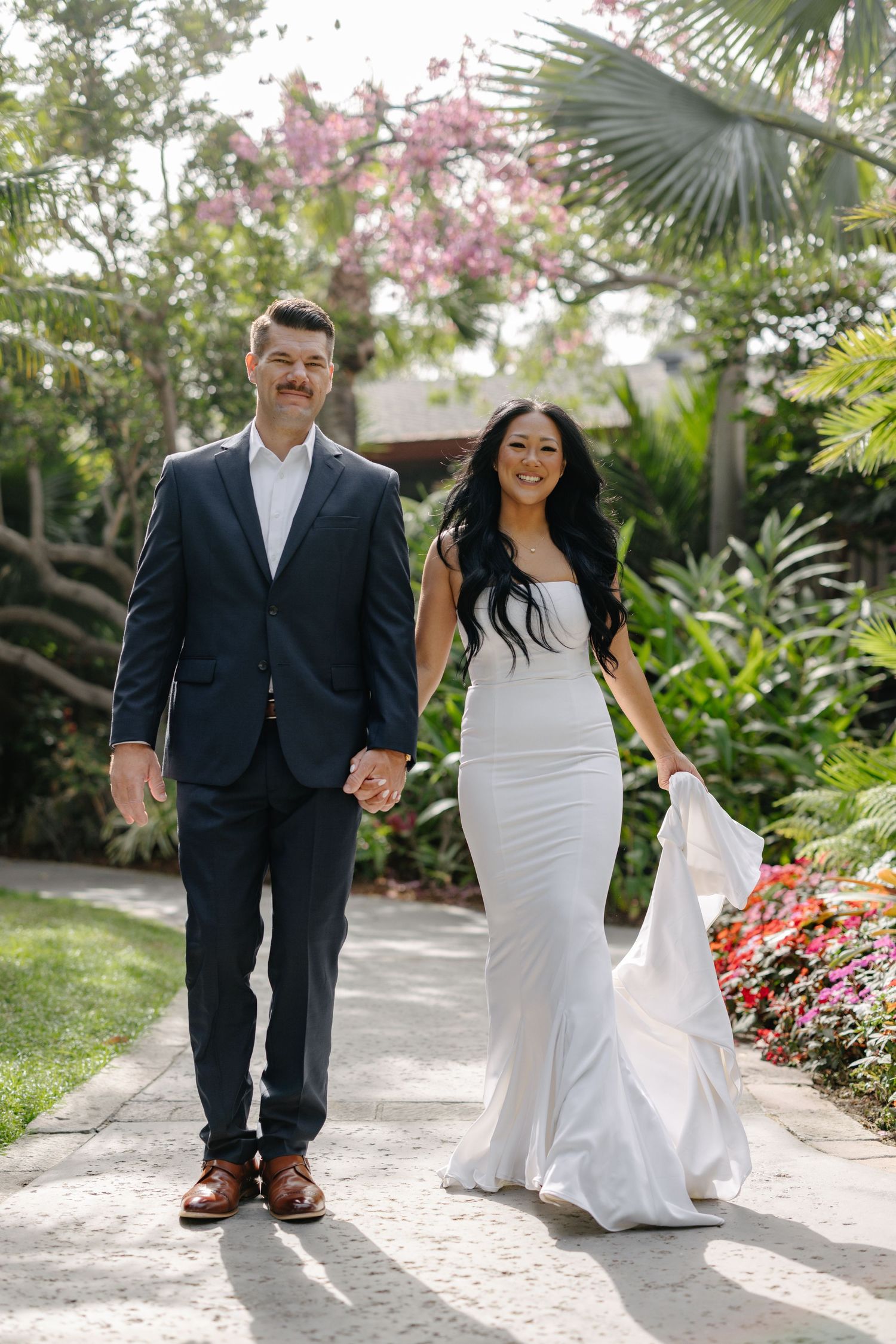 A couple in wedding attire walks hand in hand along a palm-lined garden path.
