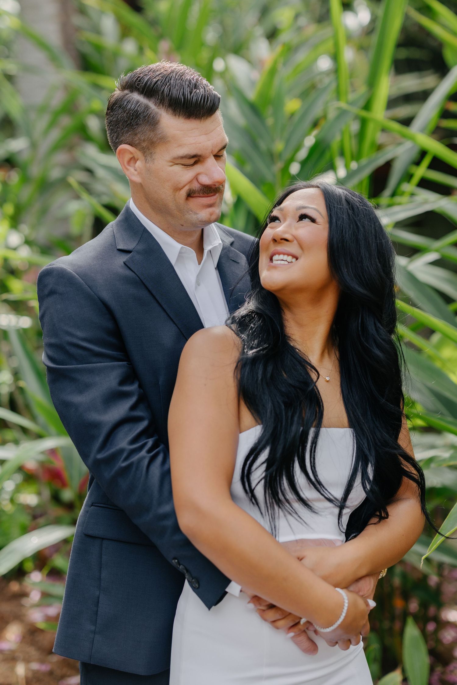 A couple in wedding attire embraces in front of tropical foliage.