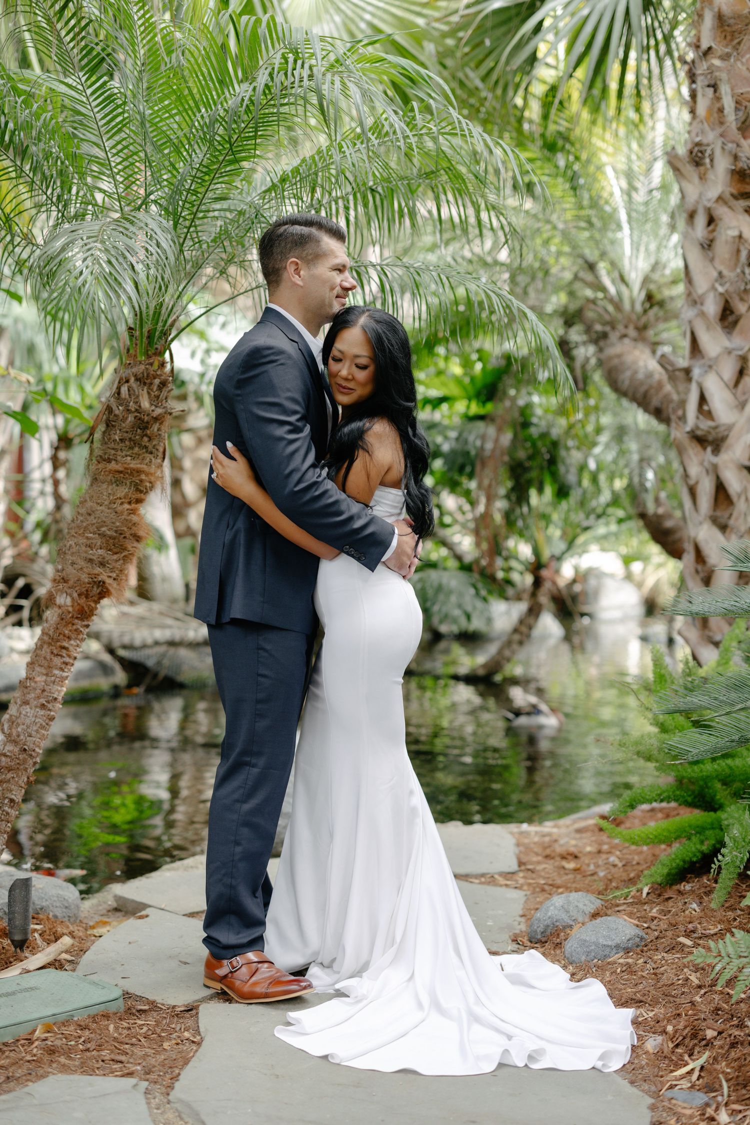 A couple embraces by a tropical pond surrounded by palm trees and lush foliage.