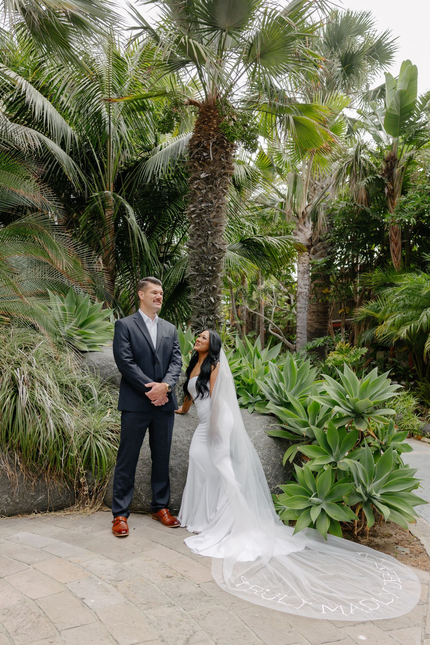 A wedding couple poses beneath towering palm trees with a flowing white veil.