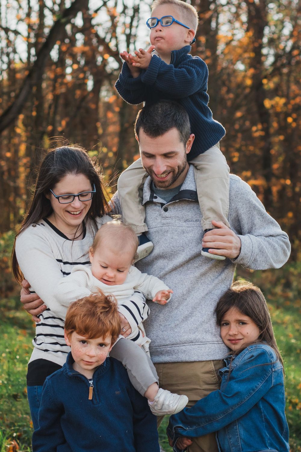 A smiling family of six poses outdoors in fall foliage, dad carrying a boy on his shoulders.