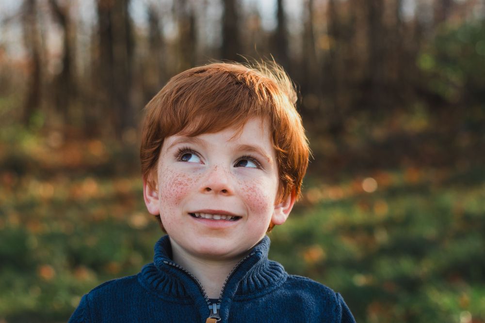 A freckled red-haired boy smiles and gazes upward in an autumn outdoor setting.