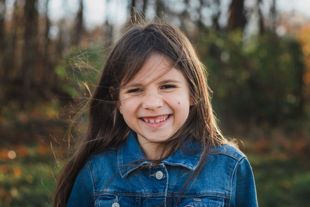 A young girl with long brown hair smiles brightly wearing a denim jacket outdoors in fall.