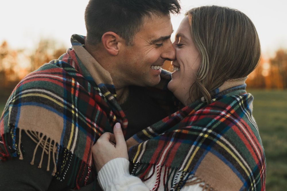 A happy couple wrapped in a plaid blanket share a joyful moment at sunset outdoors.