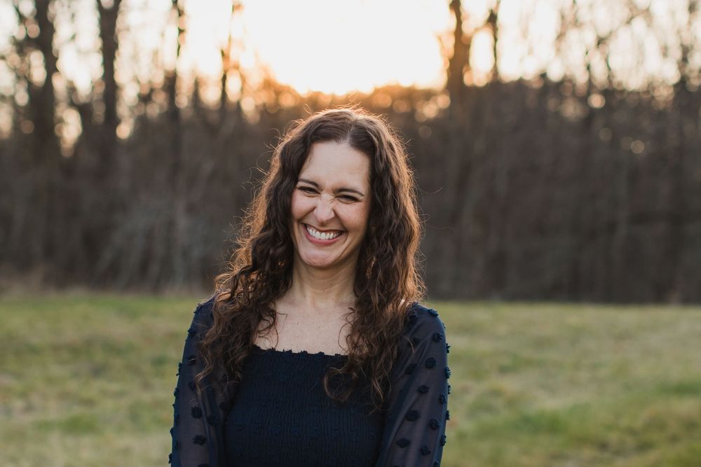 A woman with curly hair smiles warmly in a golden sunset field portrait.