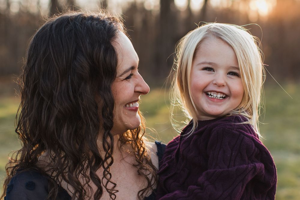A curly-haired mother smiles warmly while holding her laughing blonde toddler daughter outdoors.