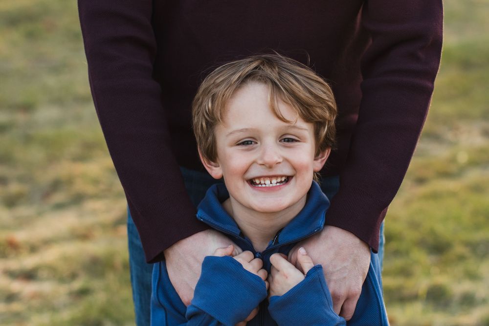 A smiling young boy in a blue jacket is hugged from behind by a parent outdoors.