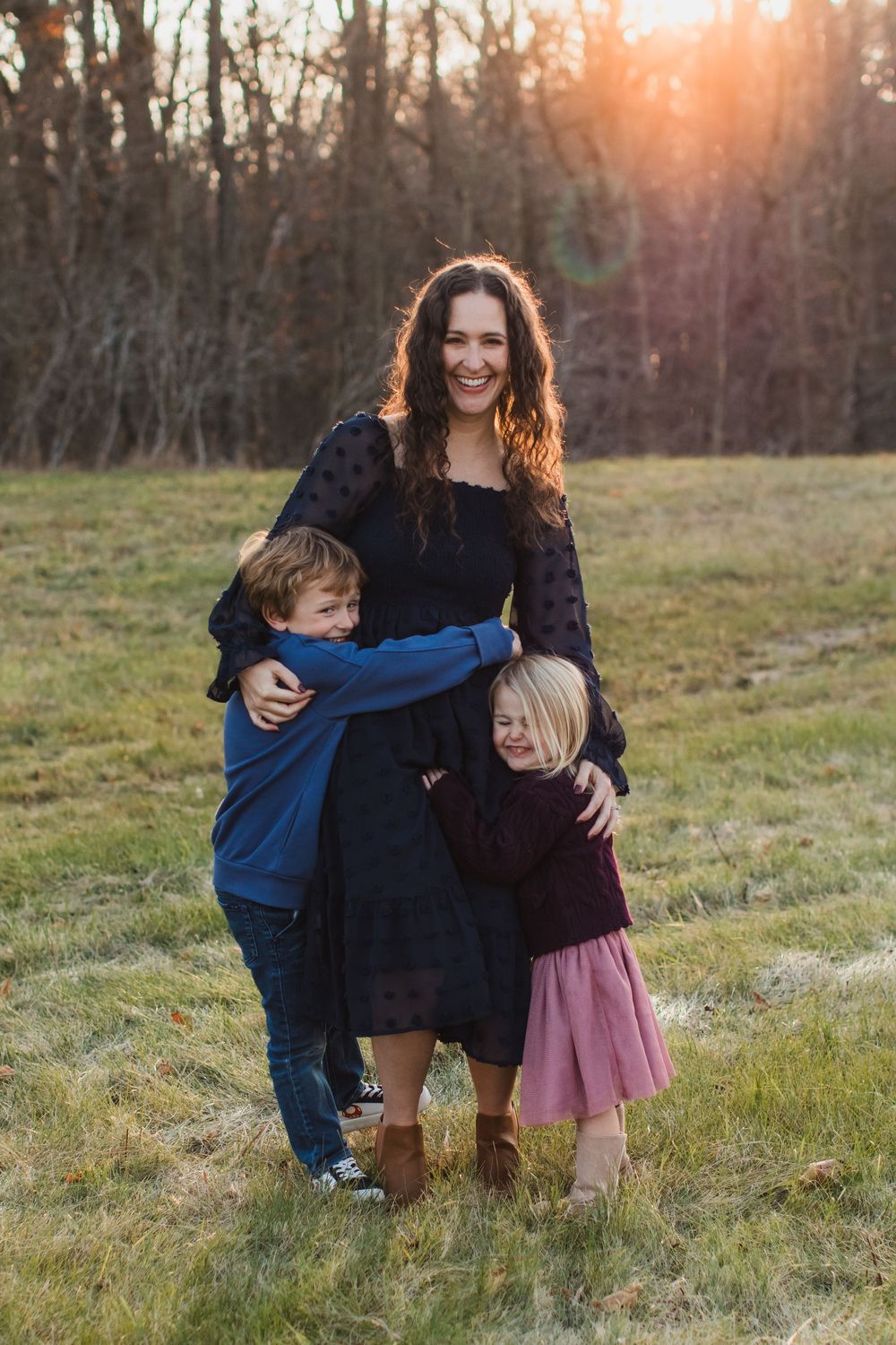 A mother in a black dress smiles as her two children hug her in a sunlit field.
