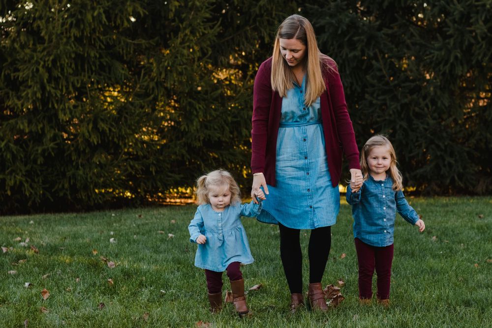 A mother walks hand-in-hand with her two young daughters in matching blue dresses on green grass.