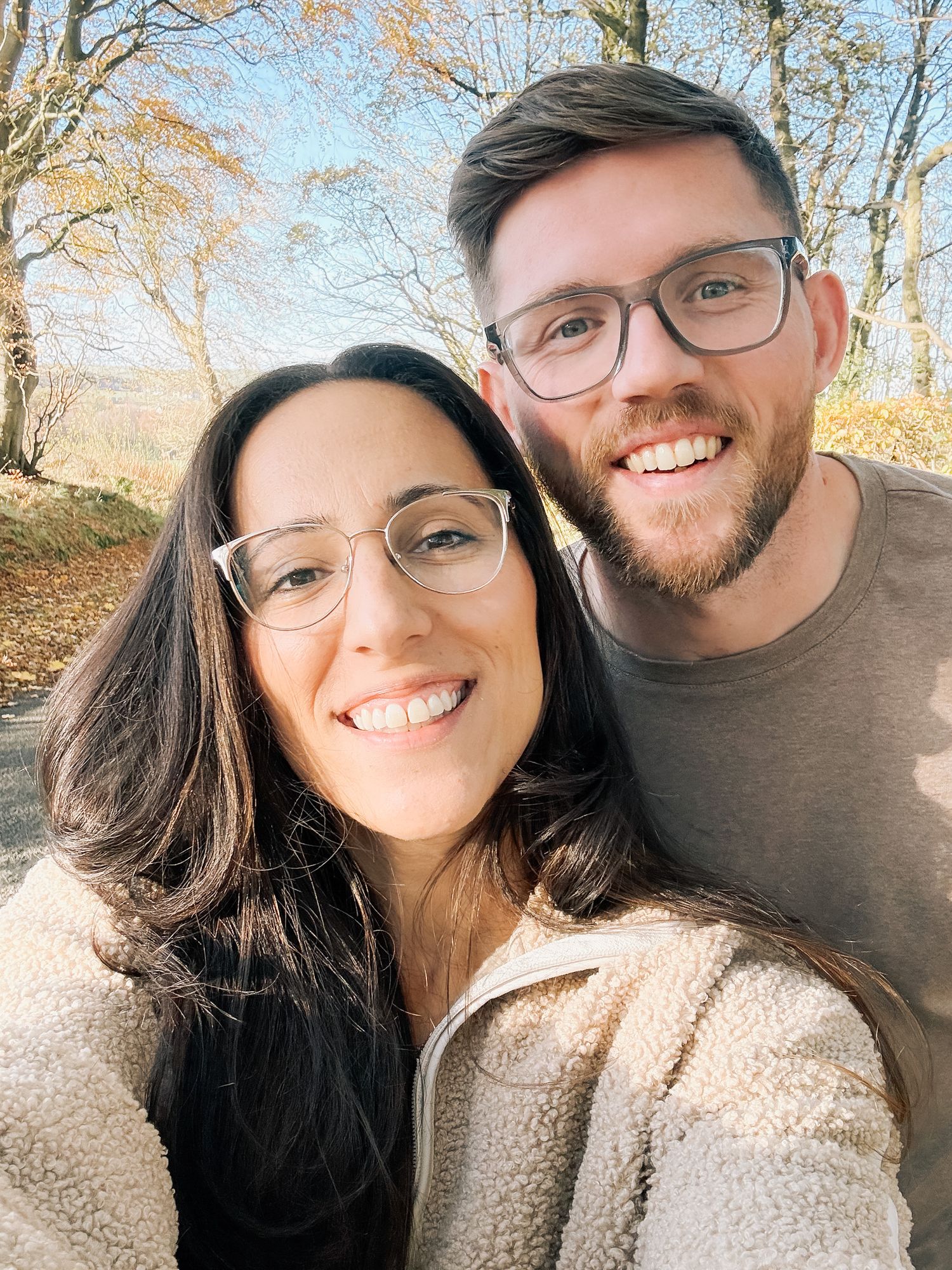 Smiling couple in glasses take a close selfie outdoors among autumn trees, wearing cozy fleece and a casual olive shirt.
