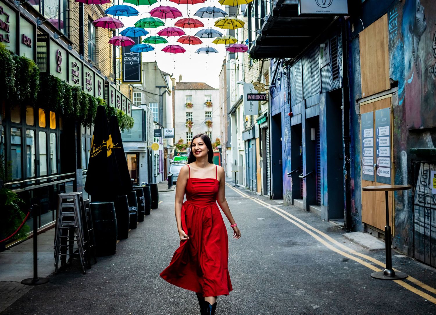 A red dress stands out against colorful umbrellas hanging above a quaint European street.