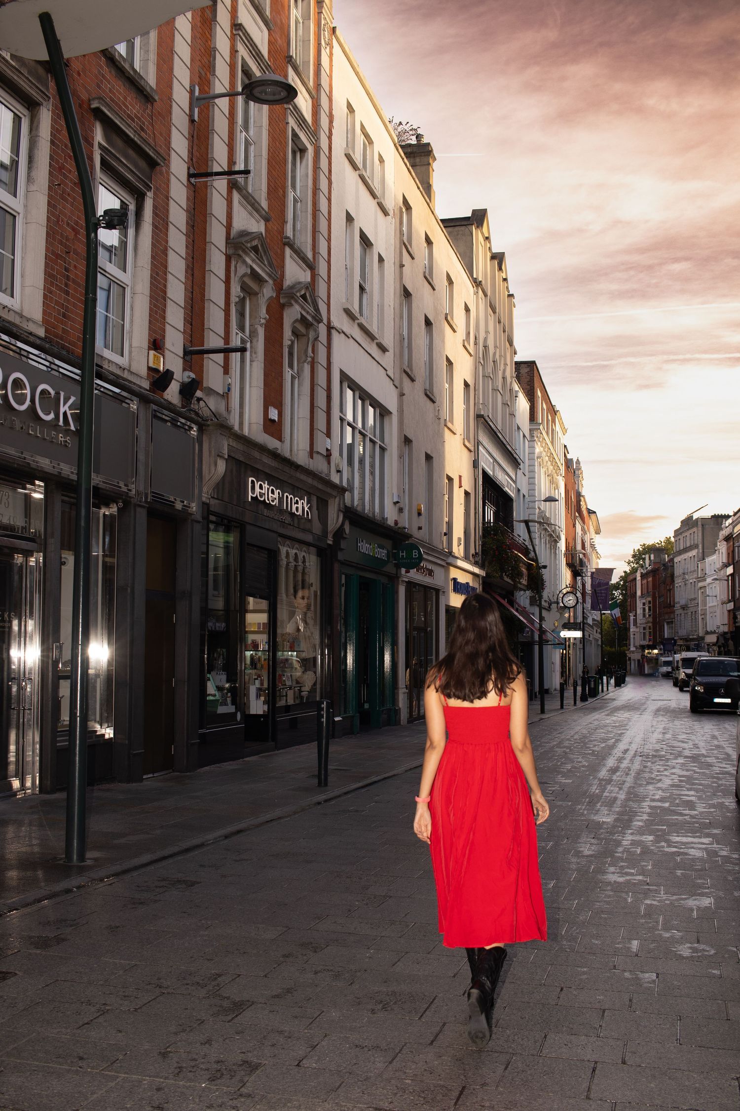 A person in a red dress walks down a dimly lit city street at dusk.