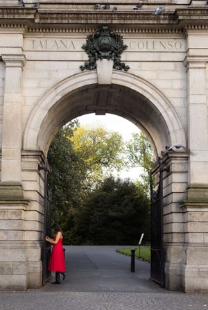 A striking red dress contrasts against a stone archway entrance to a park.