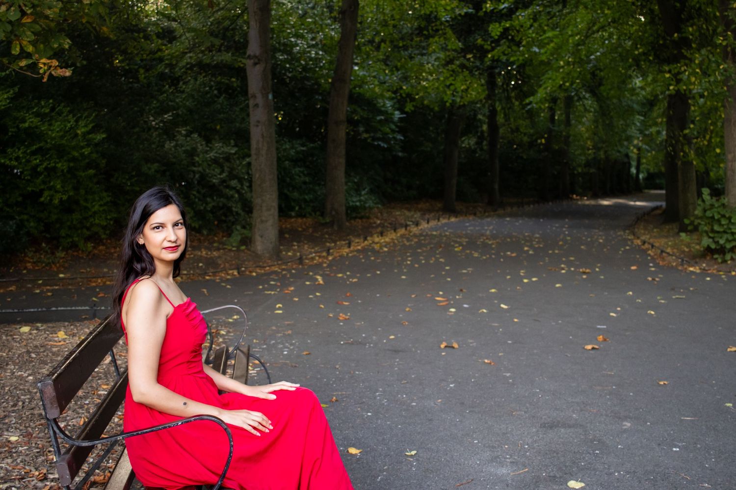 A person in a red dress sits on a park bench surrounded by autumn trees.