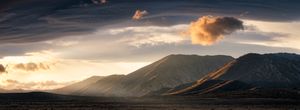 Dramatic panoramic sunset over mountain range with golden clouds and dark silhouetted peaks in a vast desert landscape.