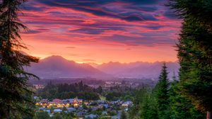 Stunning sunset over a mountain town with vibrant red and pink skies, framed by tall evergreen trees.
