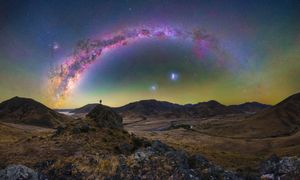 Panoramic Milky Way arch over rugged hills with a lone figure silhouetted against a vibrant starry night sky.