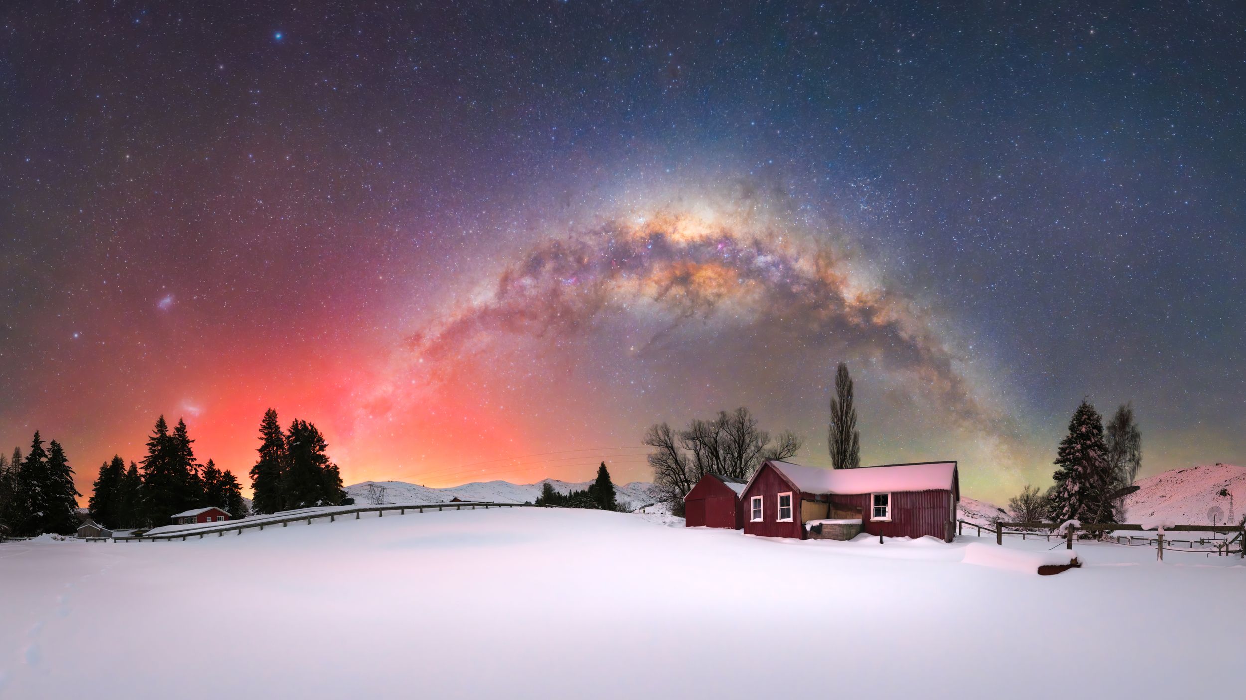 Red farmhouse under a stunning Milky Way arch with colorful aurora lights over a snowy winter landscape at night.