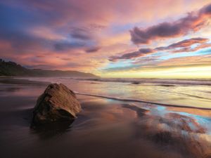 A stunning beach sunset with vibrant pink and purple clouds reflecting on wet sand beside a large rock.