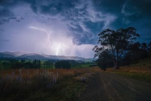 Lightning storm illuminates dramatic dark clouds over a rural dirt road with trees and mountains in the background.