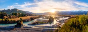 Panoramic sunset over a braided river valley with snow-capped mountains, golden light, and lush green trees in New Zealand.