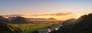 Panoramic sunset view over a lush green valley with winding river, framed by mountains and glowing orange sky.