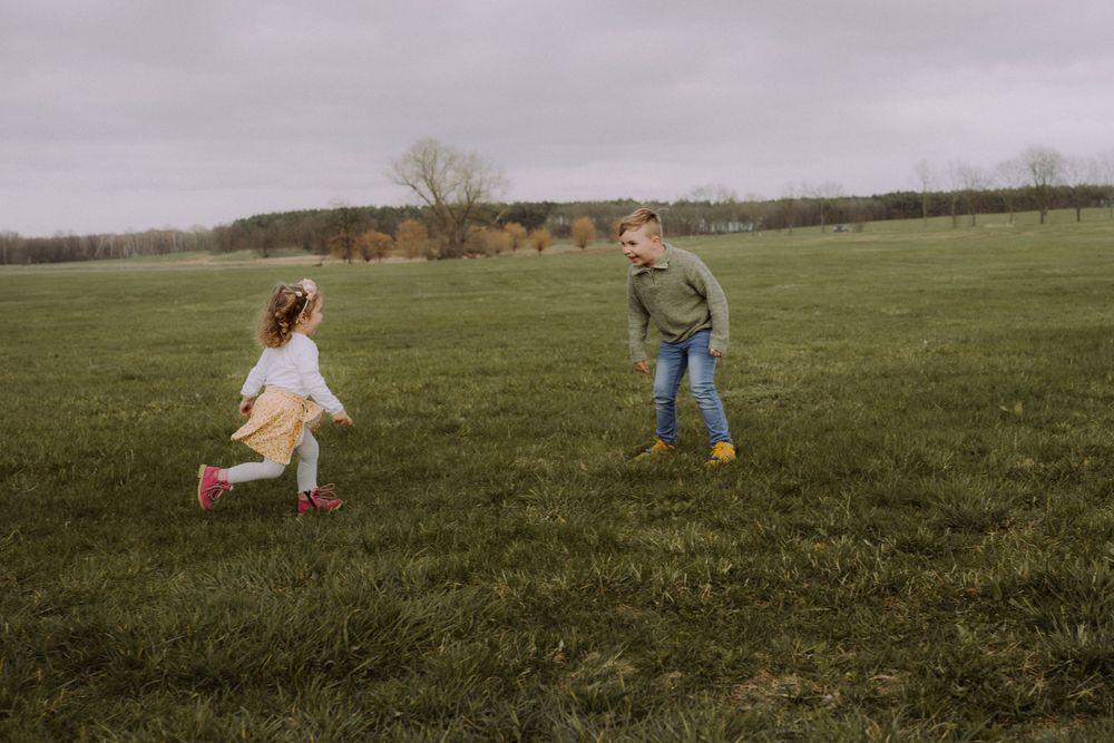 Two people run playfully through a green grassy field on an overcast autumn day.