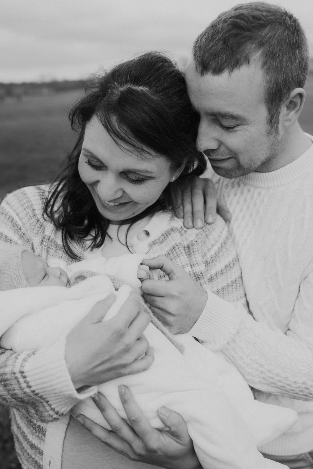 A black and white portrait of parents sharing a tender moment with their newborn baby outdoors.